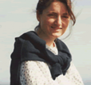 Portrait d'Elizabeth Neeser, jeune femme souriante aux cheveux bruns portant un chandail blanc et un foulard foncé, en extérieur