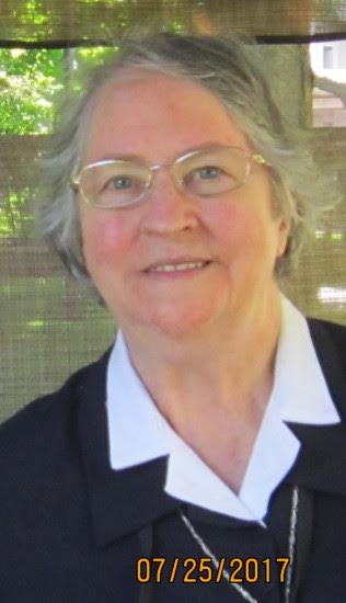 Portrait de Yolande, femme aux cheveux gris portant des lunettes et un chemisier blanc avec veste foncée, en extérieur dans un jardin