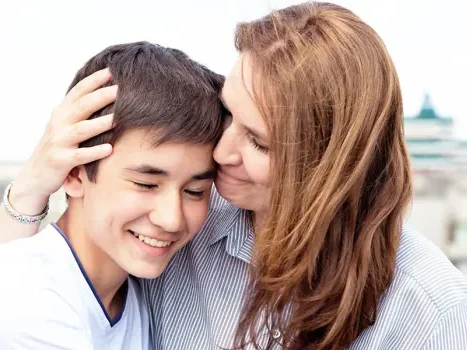 Un adolescent souriant et une femme aux cheveux roux qui pose tendrement sa tête contre la sienne en lui caressant les cheveux.