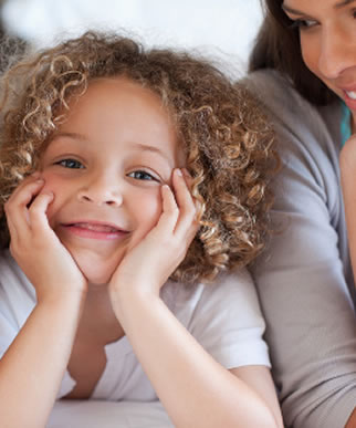 Une petite fille aux cheveux bouclés sourit à la caméra en posant son visage entre ses mains, allongée, tandis qu'une femme adulte la regarde affectueusement en arrière-plan.