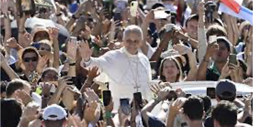 Le Pape Léon XIV salue une foule nombreuse depuis un véhicule découvert. Les personnes rassemblées lèvent les mains, prennent des photos avec leurs téléphones et accueillent chaleureusement le cortège lors d'un événement public en plein air.