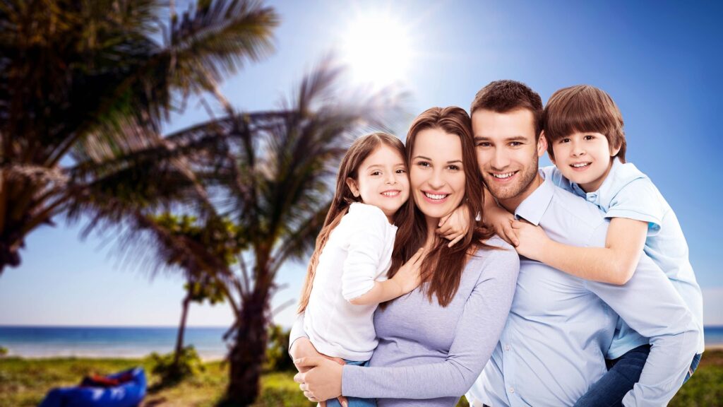 Texte alternatif :
« Une famille souriante composée de deux parents et de deux enfants pose enlacée sur une plage tropicale bordée de palmiers, sous un ciel bleu ensoleillé. »
