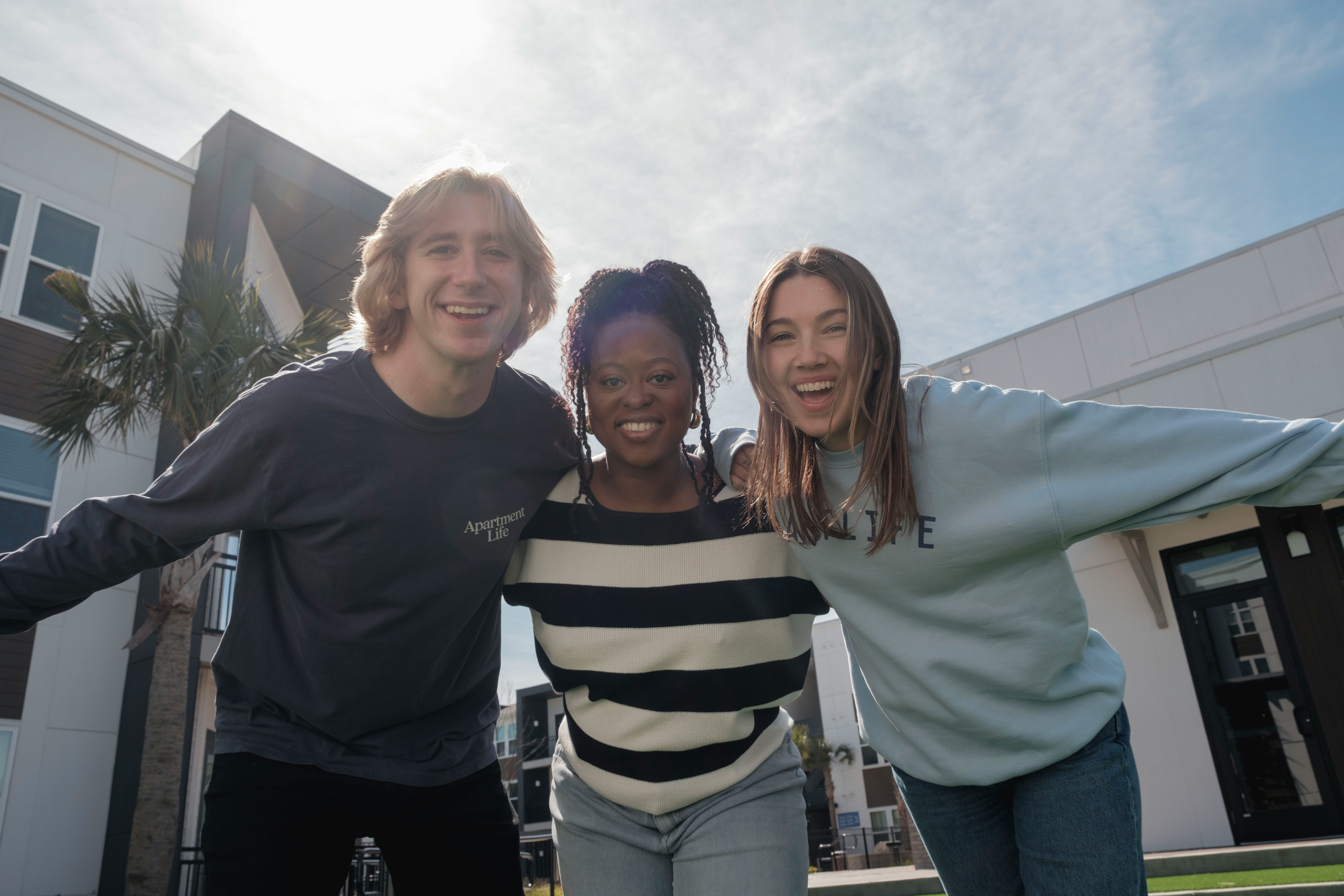 **Texte alternatif :**Trois jeunes adultes souriants se tiennent côte à côte, les bras autour des épaules, et regardent l'appareil photo dans une cour extérieure devant un immeuble résidentiel moderne. Le ciel est dégagé et ensoleillé, avec quelques palmiers et des bâtiments en arrière-plan, créant une ambiance conviviale et chaleureuse.