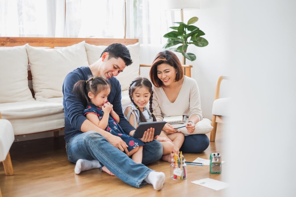 Famille assise au sol dans un salon lumineux, deux parents et leurs deux jeunes filles regardant une tablette ensemble, avec des crayons et un carnet à proximité.