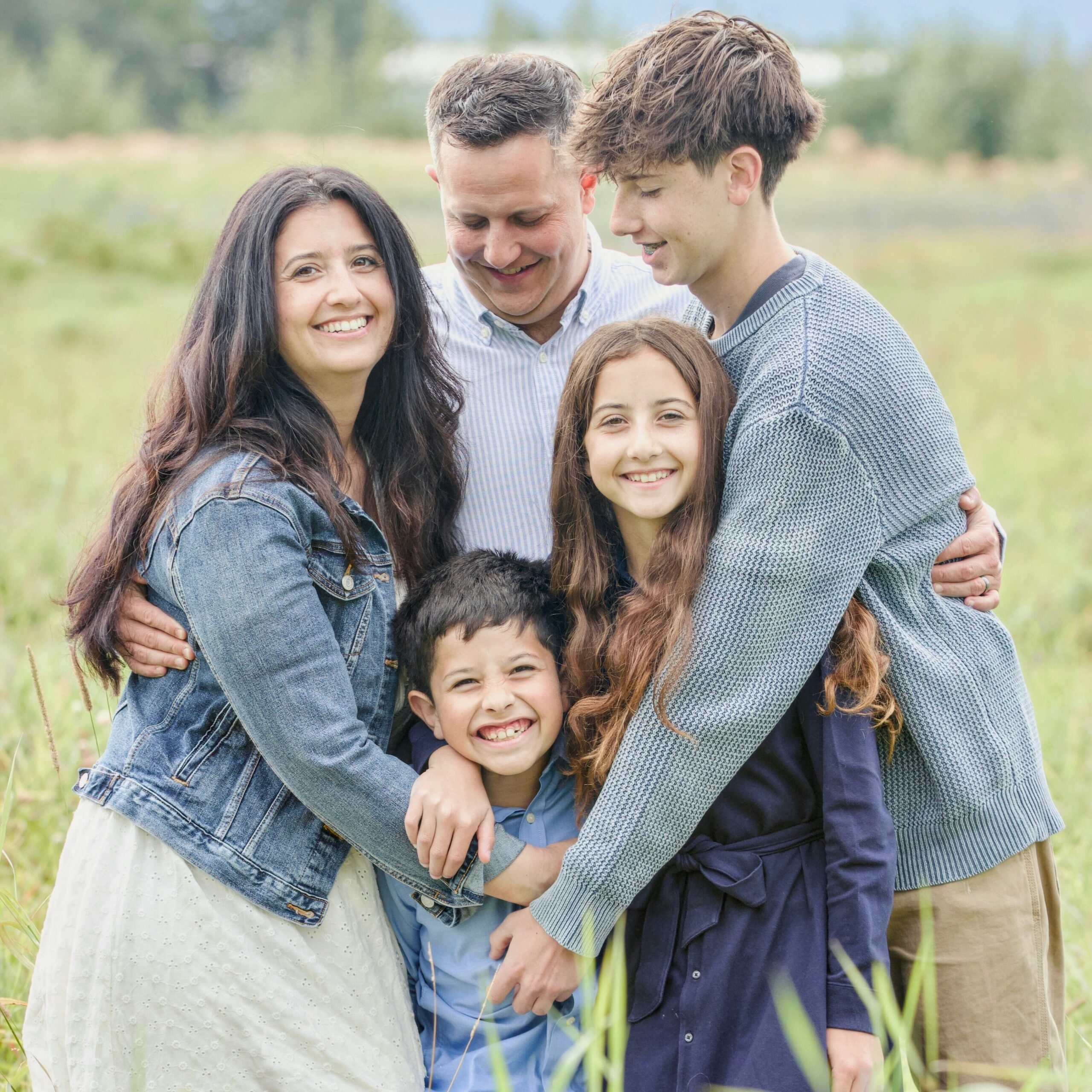 Une famille de cinq personnes sourit et se serre affectueusement dans les bras, debout dans une prairie verdoyante. On y voit deux parents et trois enfants — un adolescent, une fillette et un jeune garçon au centre qui rit aux éclats — avec en arrière-plan des arbres.